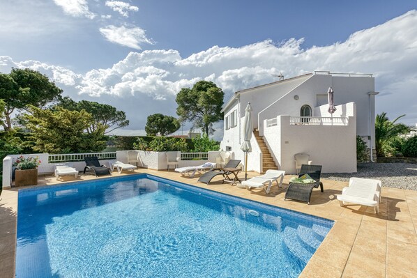 A swimming pool with lounge chairs, a white building with a balcony, and trees in the background.