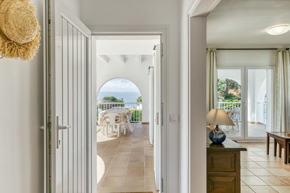An open door leading to a balcony with a table and chairs, a view of the sea, and a lamp on a dresser.