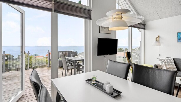 A modern room with a large window overlooking the sea, a dining area with a table and chairs, a mounted TV, and a ceiling light fixture. - Lovely home in Esbjerg V with WiFi (Esbjerg V)