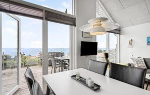 Une chambre moderne dotée d’une grande fenêtre donnant sur la mer, d’un coin repas avec une table et des chaises, d’un téléviseur fixé au mur et d’un luminaire suspendu au plafond.