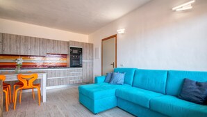 Una cocina moderna con horno y microondas integrados, una zona de comedor con sillas anaranjadas y un sofá color turquesa.