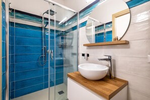 A modern bathroom with a glass-enclosed shower, a white sink, and a mirror with a wooden shelf.