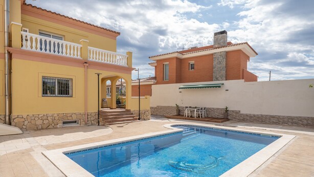 Una piscina con borde de piedra, un edificio amarillo con balcón y un edificio de ladrillo rojo al fondo.