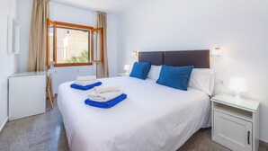 A hotel room with a bed, a desk, a chair, a window with curtains, a nightstand with a lamp, and a wall-mounted light. - Apartment 'Apartamento Cabrera' with Private Terrace, Wi-Fi and Air Conditioning (Colònia de Sant Jordi)