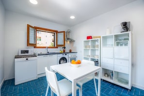 Une cuisine compacte équipée d’un four à micro-ondes, d’un lave-linge et d’un évier. On y trouve une table à manger avec des chaises et un saladier rempli de fruits.