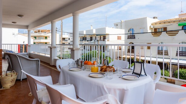 A balcony with white tables and chairs, a breakfast setting with plates, glasses, and orange juice, and a view of buildings in the background. - Apartment 'Apartamento Cabrera' with Private Terrace, Wi-Fi and Air Conditioning (Colònia de Sant Jordi)