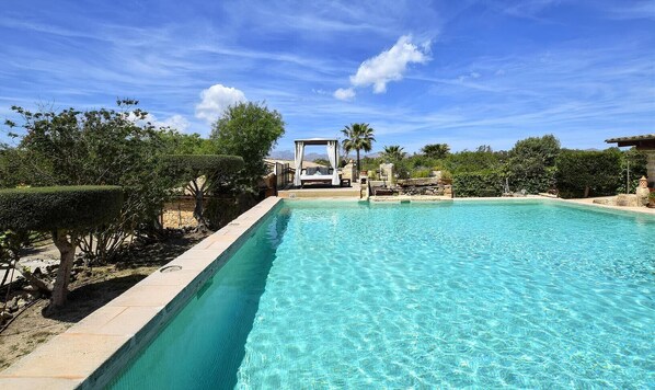 Una piscina rettangolare con acqua cristallina e blu, circondata da vegetazione e un muro in pietra.