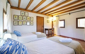 A room with two beds, a wooden ceiling, a desk with a chair, and framed pictures on the wall.