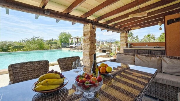 Een terras met rieten meubilair, een tafel met fruit en wijn, en uitzicht op een zwembad en bergen.