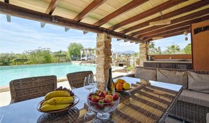 Zona pranzo all'aperto con mobili in vimini, un tavolo con frutta e vino, e vista su piscina e montagne.