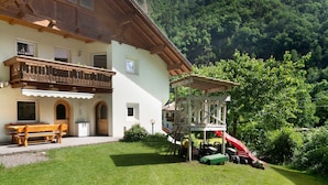 A chalet with a balcony, a wooden bench, a slide, and a mountain backdrop.