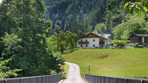 A rural road leading to a white building with a balcony, surrounded by greenery and hills.