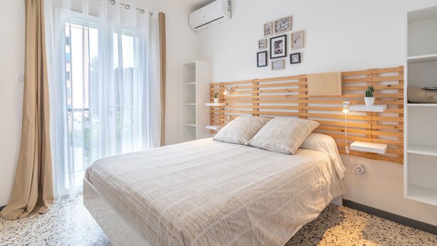 A bedroom with a bed, a window with sheer curtains, a wooden headboard, wall-mounted shelves, and a wall-mounted air conditioner. - Canigou Et Jo room with shared garden, Wi-Fi and air conditioning (Perpignan)