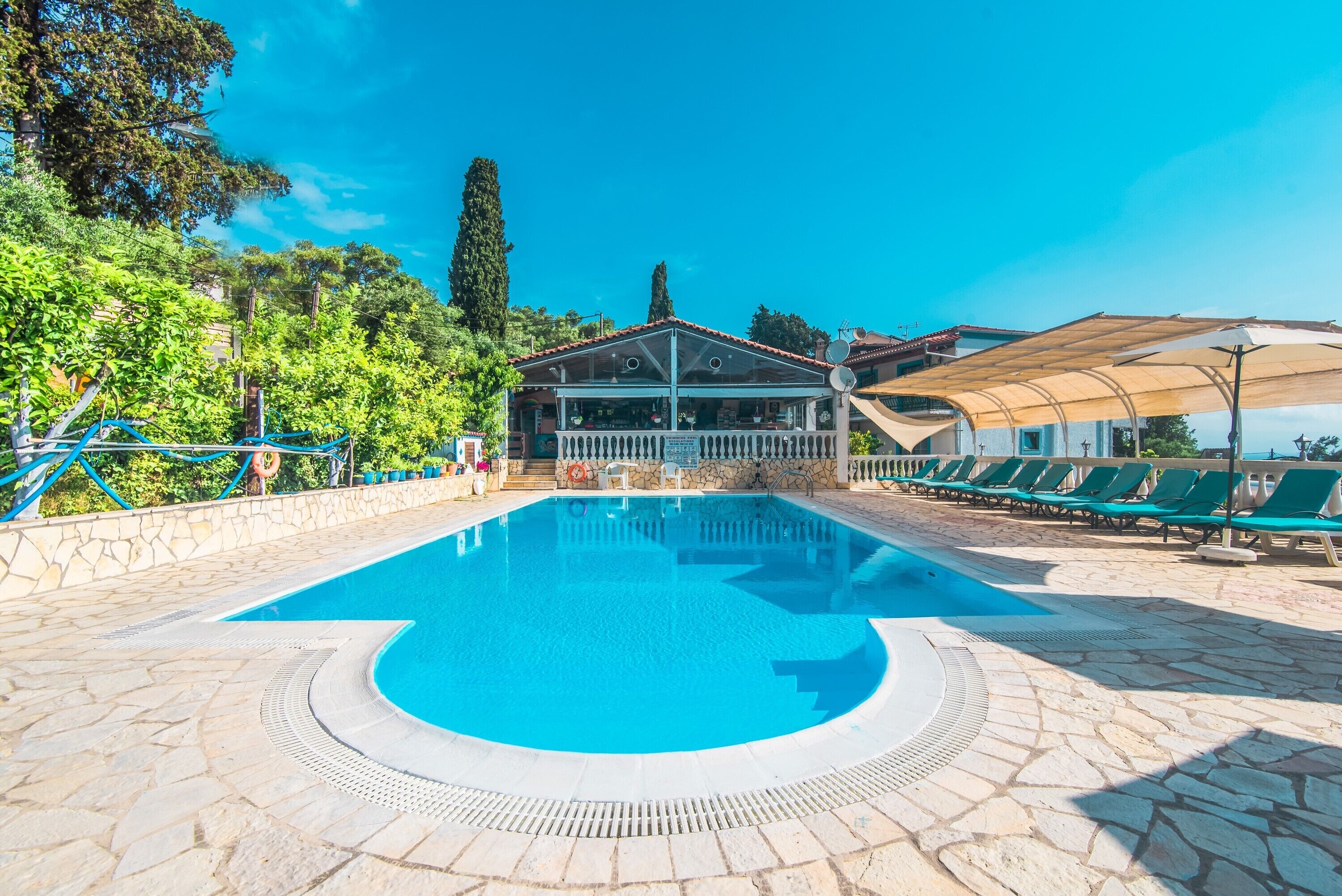 A swimming pool with a stone deck, a covered seating area, and a building with a tiled roof in the background.