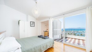 Een slaapkamer met een bed, een nachtkastje, een hanglamp, en uitzicht op een jachthaven door een openslaande balkondeur, en een geruite vloer.