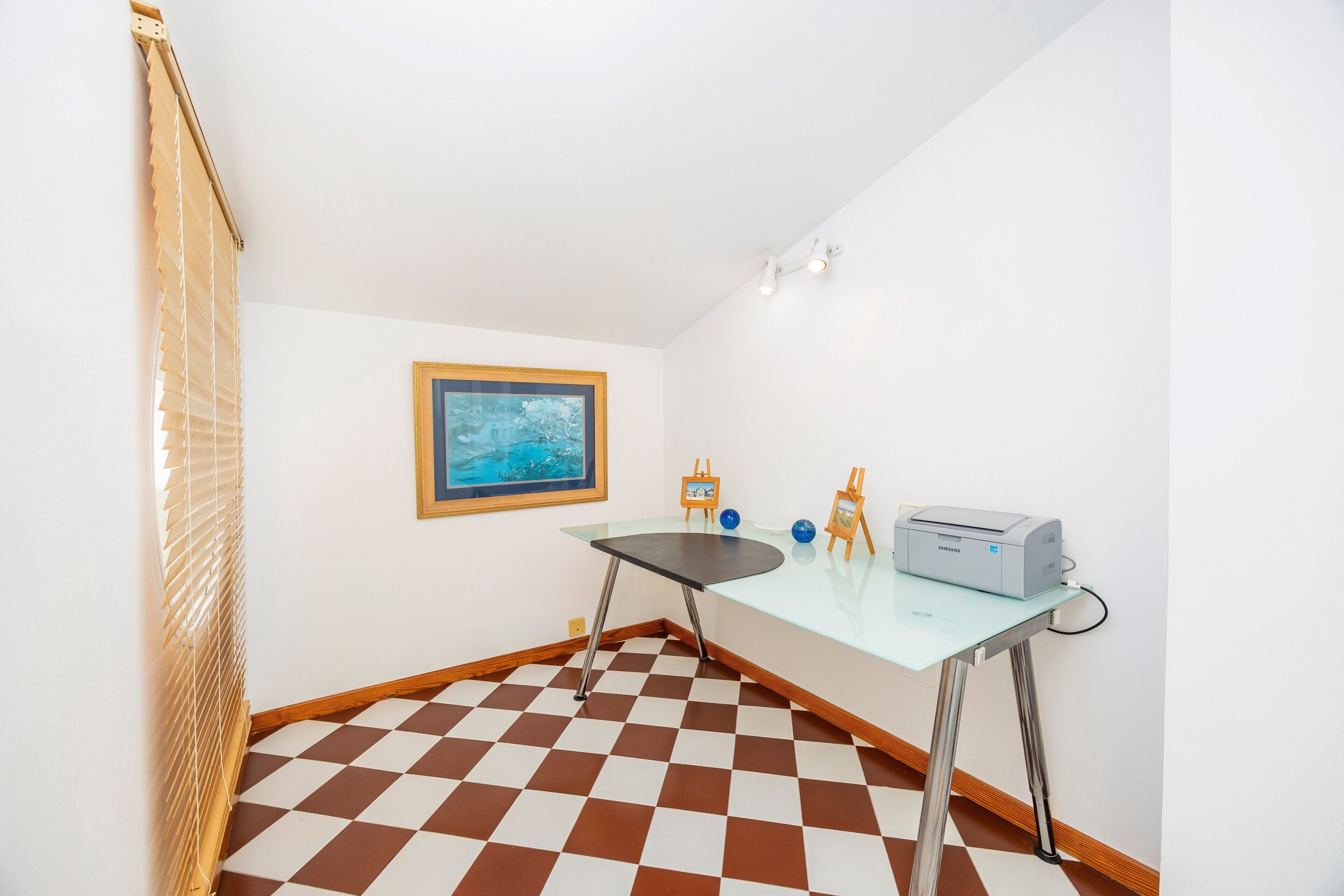 A room with a checkered floor, a glass table with a printer, a framed picture, and a window with blinds.
