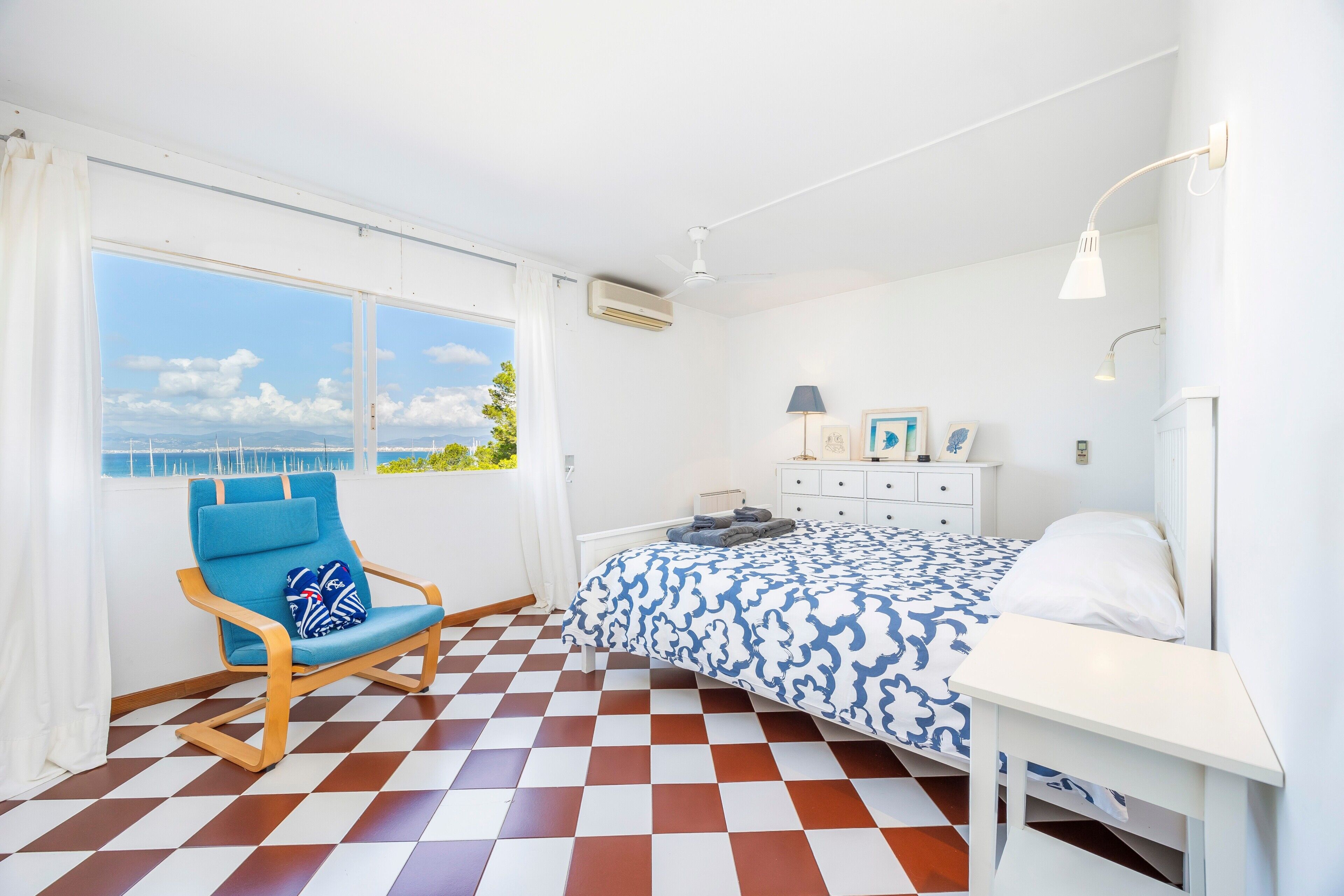 A bedroom with a checkered floor, a bed with a patterned bedspread, a rocking chair, a bedside table, and a window with a view of the sea.