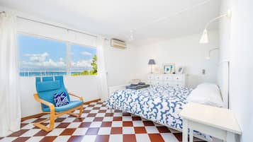 A bedroom with a checkered floor, a bed with a patterned bedspread, a rocking chair, a bedside table, and a window with a view of the sea.