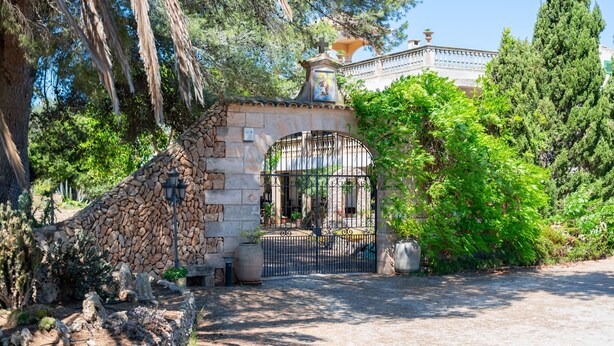 Un arco de piedra que da acceso a una entrada con reja, rodeado de follaje y árboles.