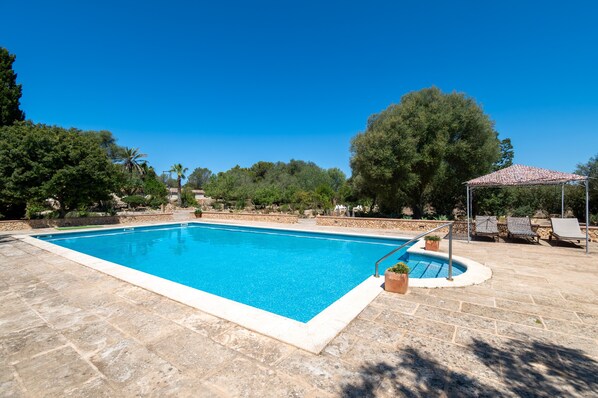 Una piscina rectangular con solárium empedrado, rodeada de árboles y una pérgola con sillas.
