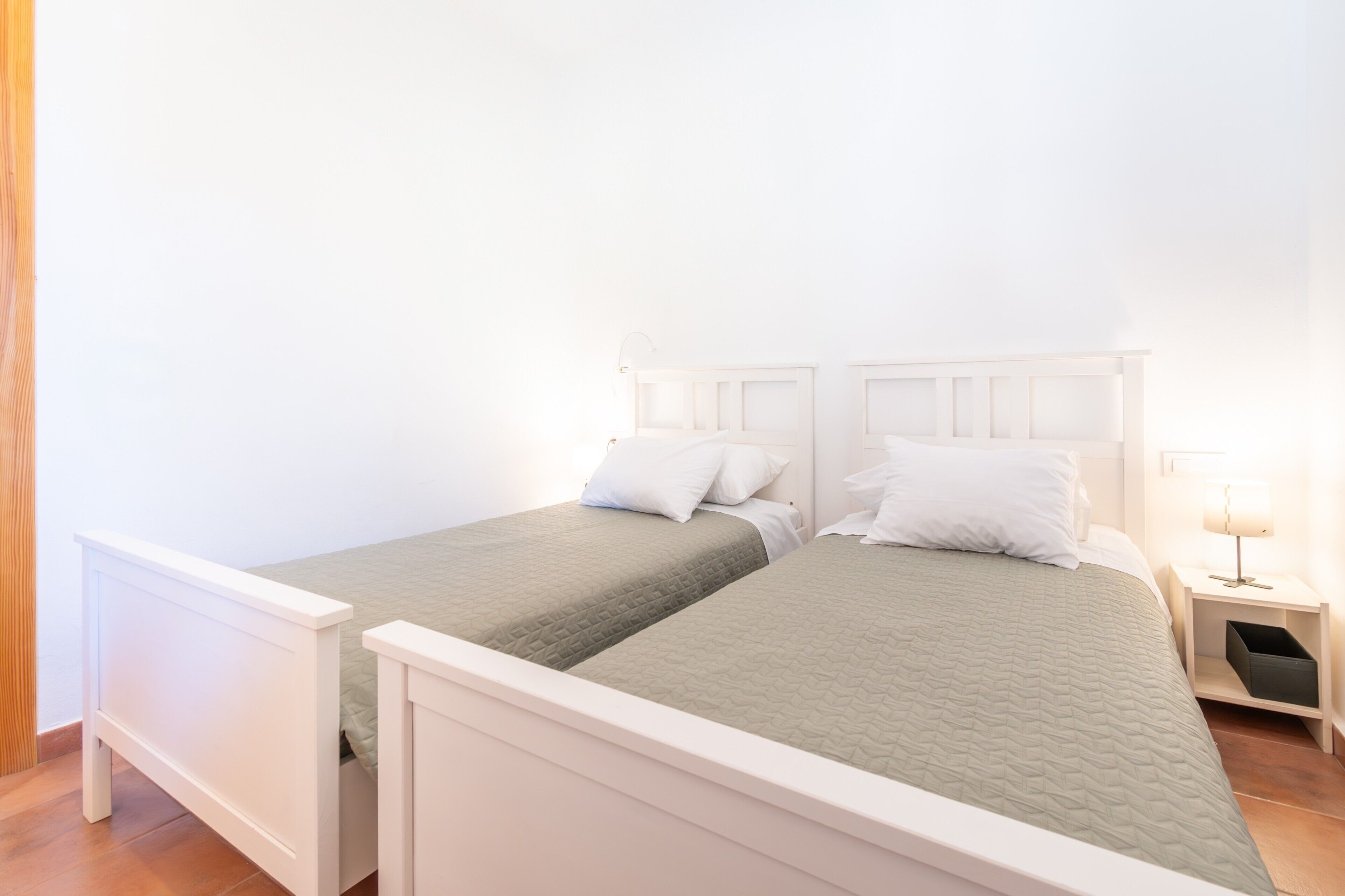 Uma cama de casal com estrutura de madeira e colcha verde, uma mesinha de cabeceira com abajur e um criado-mudo com caixa de armazenamento.