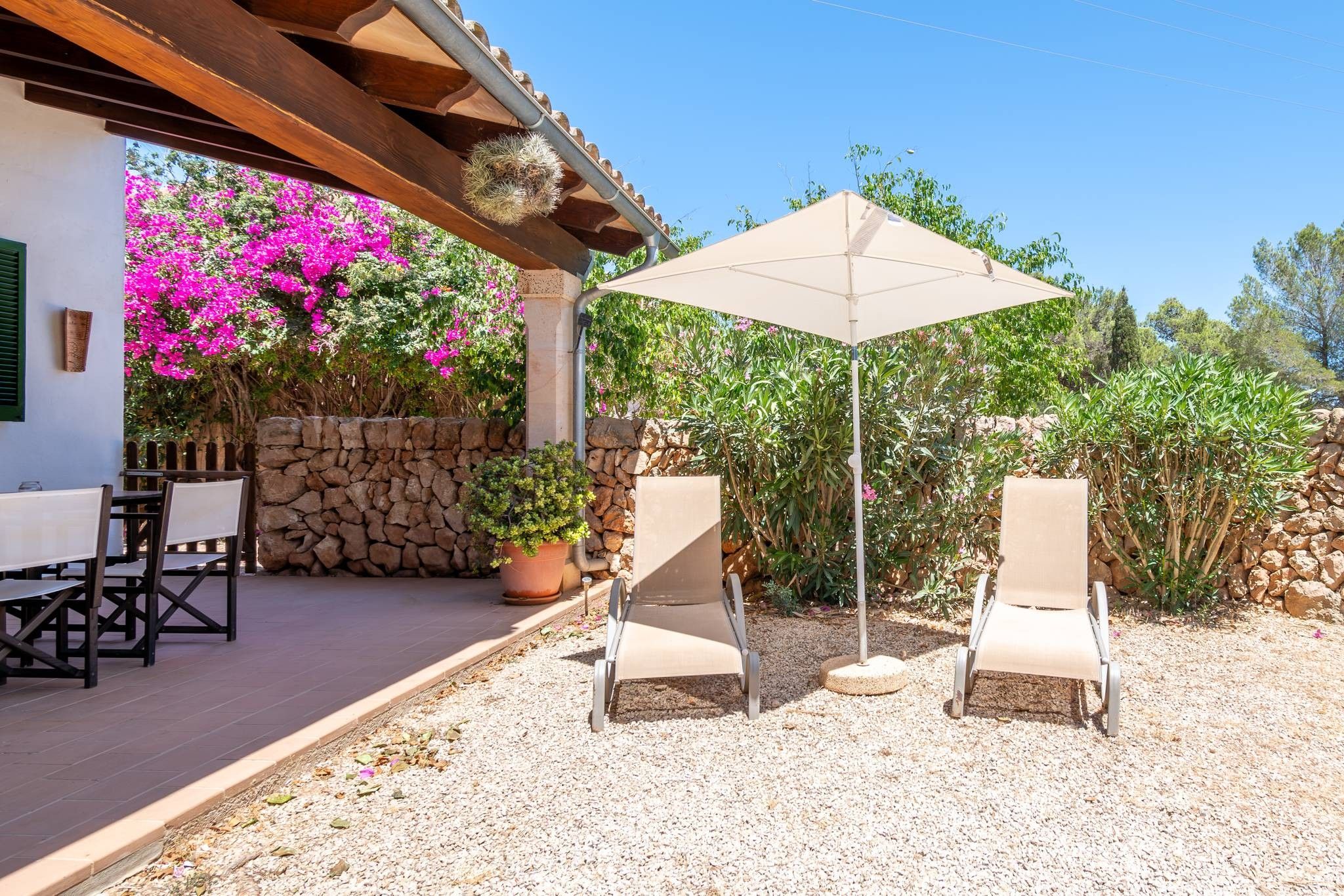 Een terras met twee stoelen en een tafel, een pergola en een stenen muur begroeid met beplanting.