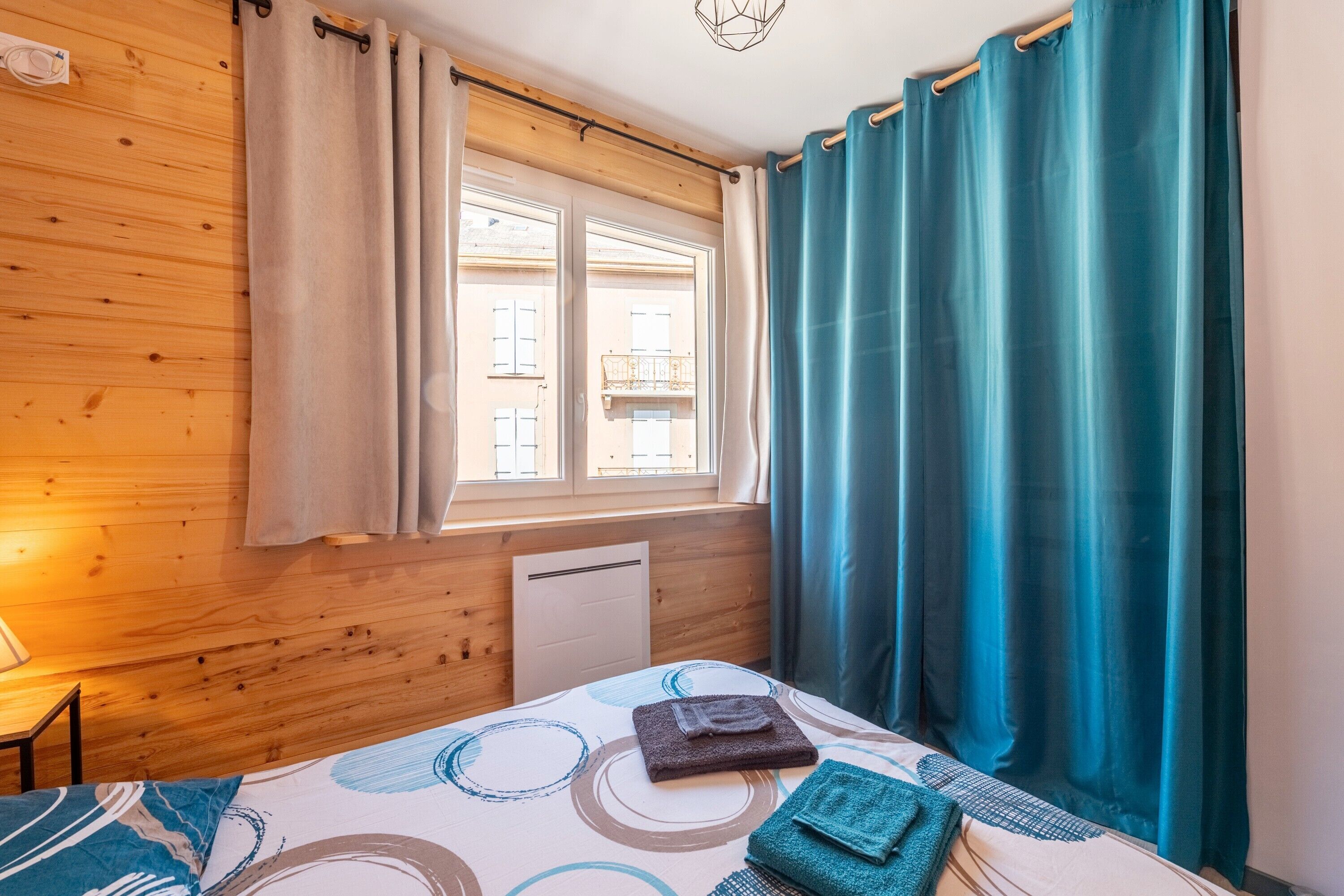 Ein Zimmer mit Holzwänden, einem Bett mit gemustertem Überwurf, einem Fenster mit Vorhängen und einem Heizkörper.
