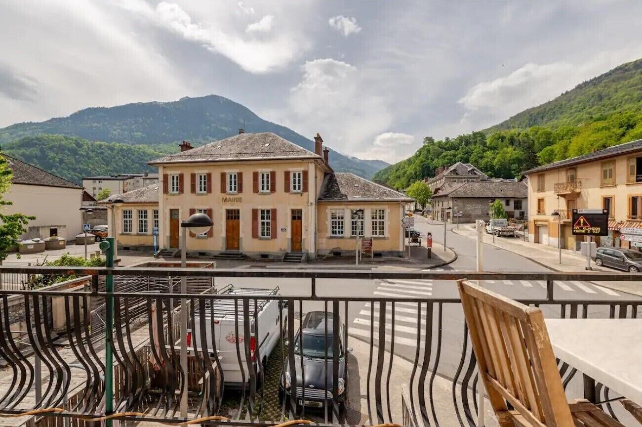 Ein Gebäude mit dem Schriftzug 'MAIRIE', eine Straße mit parkenden Autos und ein Berg im Hintergrund.