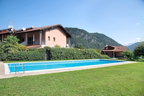 Ein zweistöckiges Haus mit Schwimmbad, umgeben von Grünflächen und Bergen.