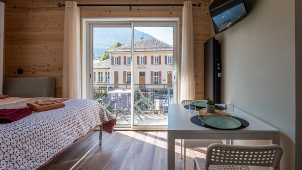 A hotel room with a bed, a table with plates, a chair, a TV, and a balcony with a view of buildings. - Apartment 'Le Chaussy' with Mountain View, Balcony and Wi-Fi (Saint-Etienne-de-Cuines)