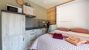 A bedroom with a bed, pillows, a blanket, a wooden wall, a window, and a kitchenette. - Apartment 'Le Chaussy' with Mountain View, Balcony and Wi-Fi (Saint-Etienne-de-Cuines)