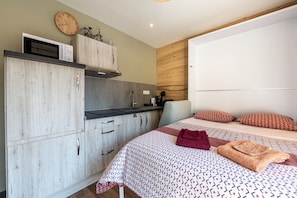 Un dormitorio con cama, almohadas, manta, pared de madera, ventana y kitchenette.