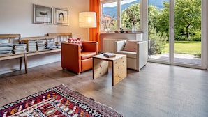 A modern living room with a wooden floor, a sofa, a chair, a coffee table, and a view of a garden through large windows.