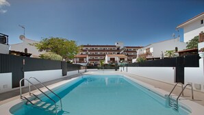 Una piscina rectangular con escaleras metálicas a ambos lados, rodeada por una valla baja, y un edificio de varios pisos al fondo.