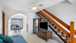 Una sala de estar con televisor, escalera de madera y comedor al fondo.