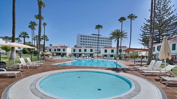 Zona de piscina de hotel con sillones, palmeras y un edificio al fondo.