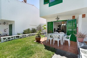 Une terrasse avec des chaises et une table en plastique blanc, un pot de fleurs et une porte verte.