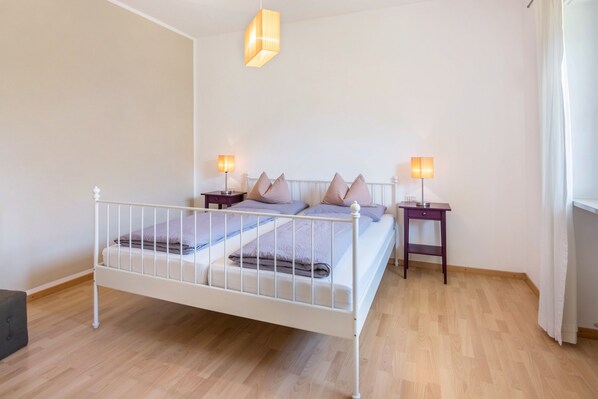 A bedroom with a white metal bed, two bedside tables with lamps, a wooden floor, and a window with curtains.