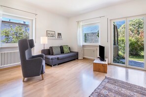 En moderne stue med en grå lænestol, en grå sofa, et tv på et træstel og store vinduer med udsigt til grøn vegetation og en stenvæg.