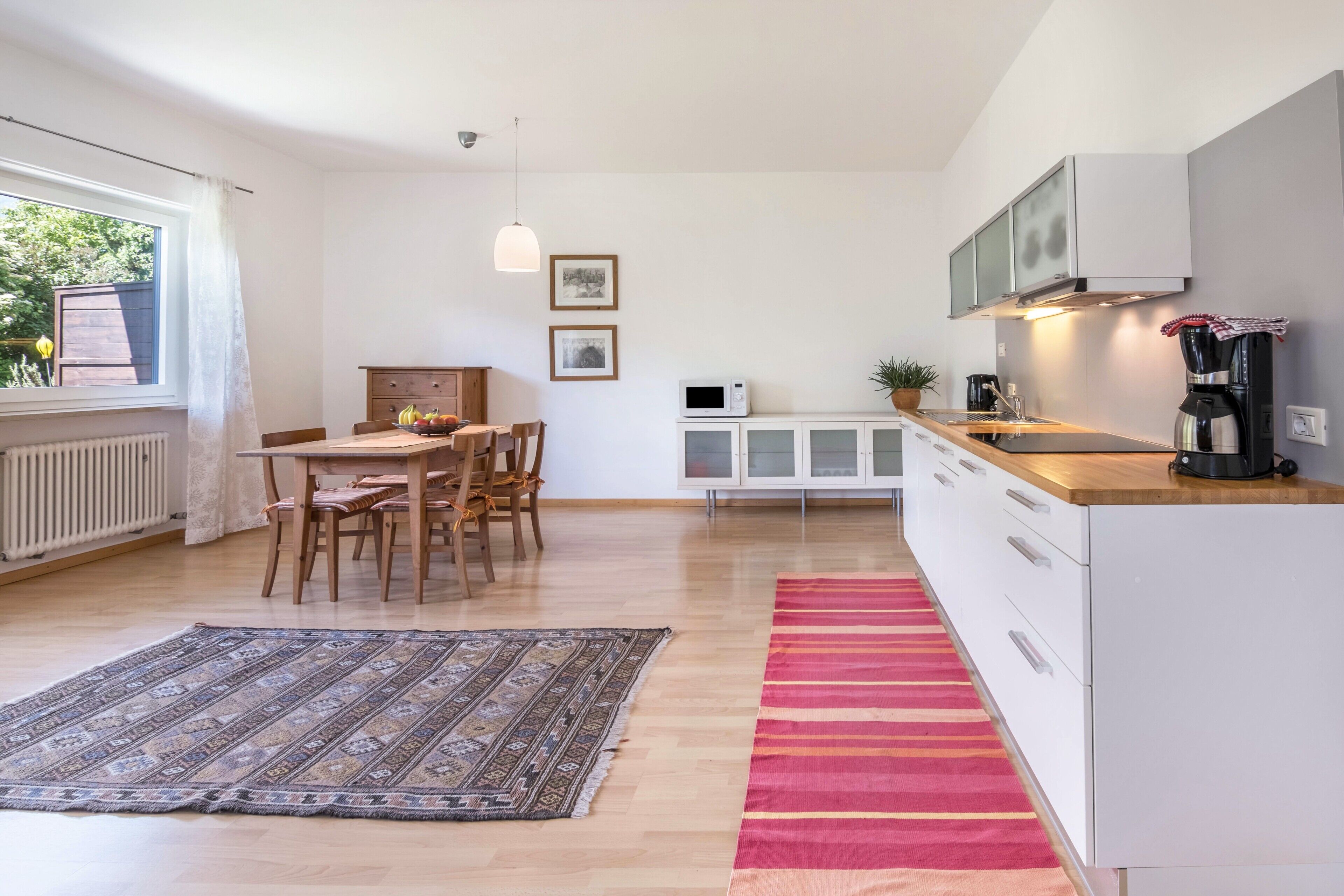 Une cuisine moderne avec des meubles blancs, un coin repas doté d’une table et de chaises en bois, et une grande fenêtre offrant une vue sur la verdure.