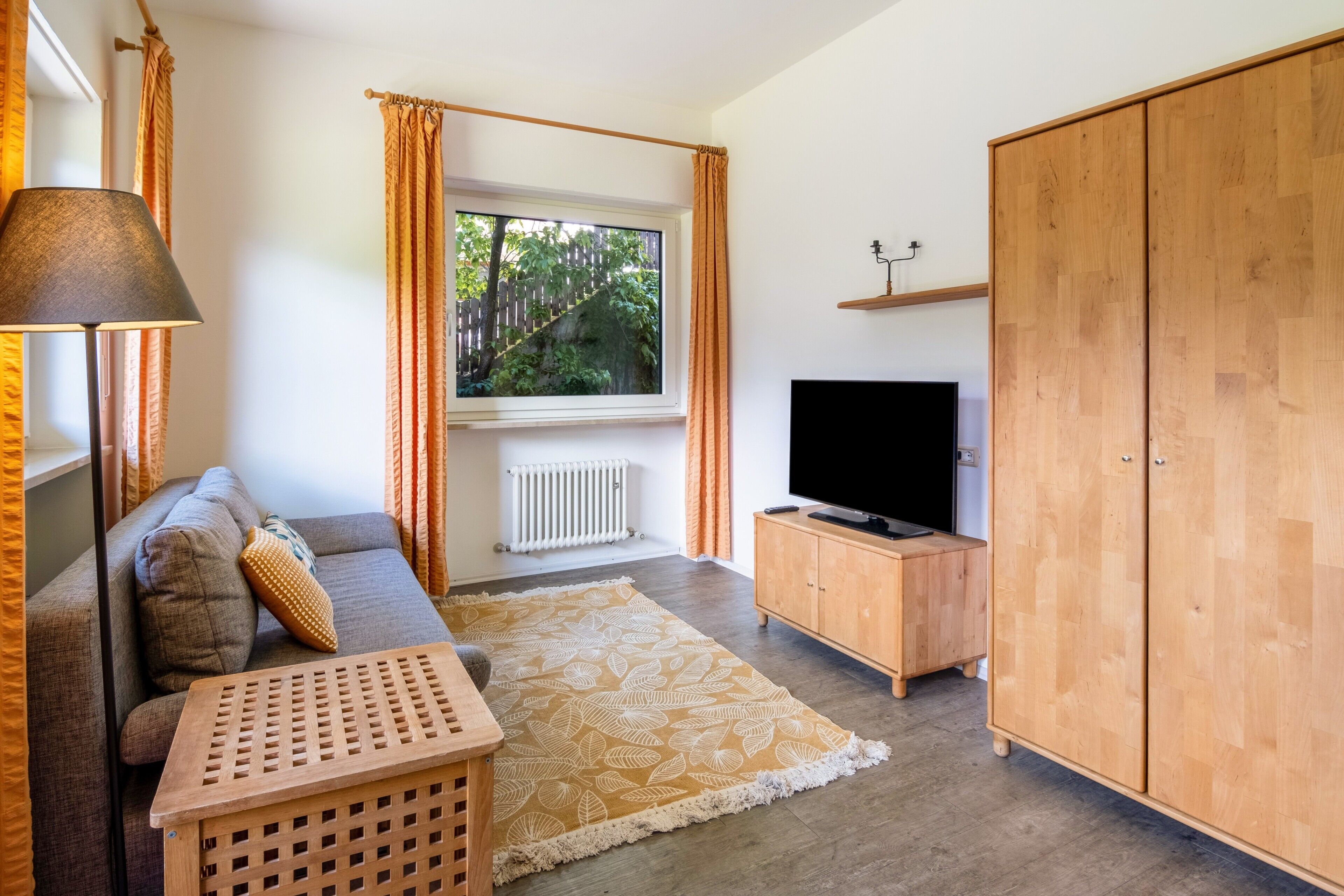 A living room with a sofa, a TV, wooden furniture, a window with curtains, and a radiator.