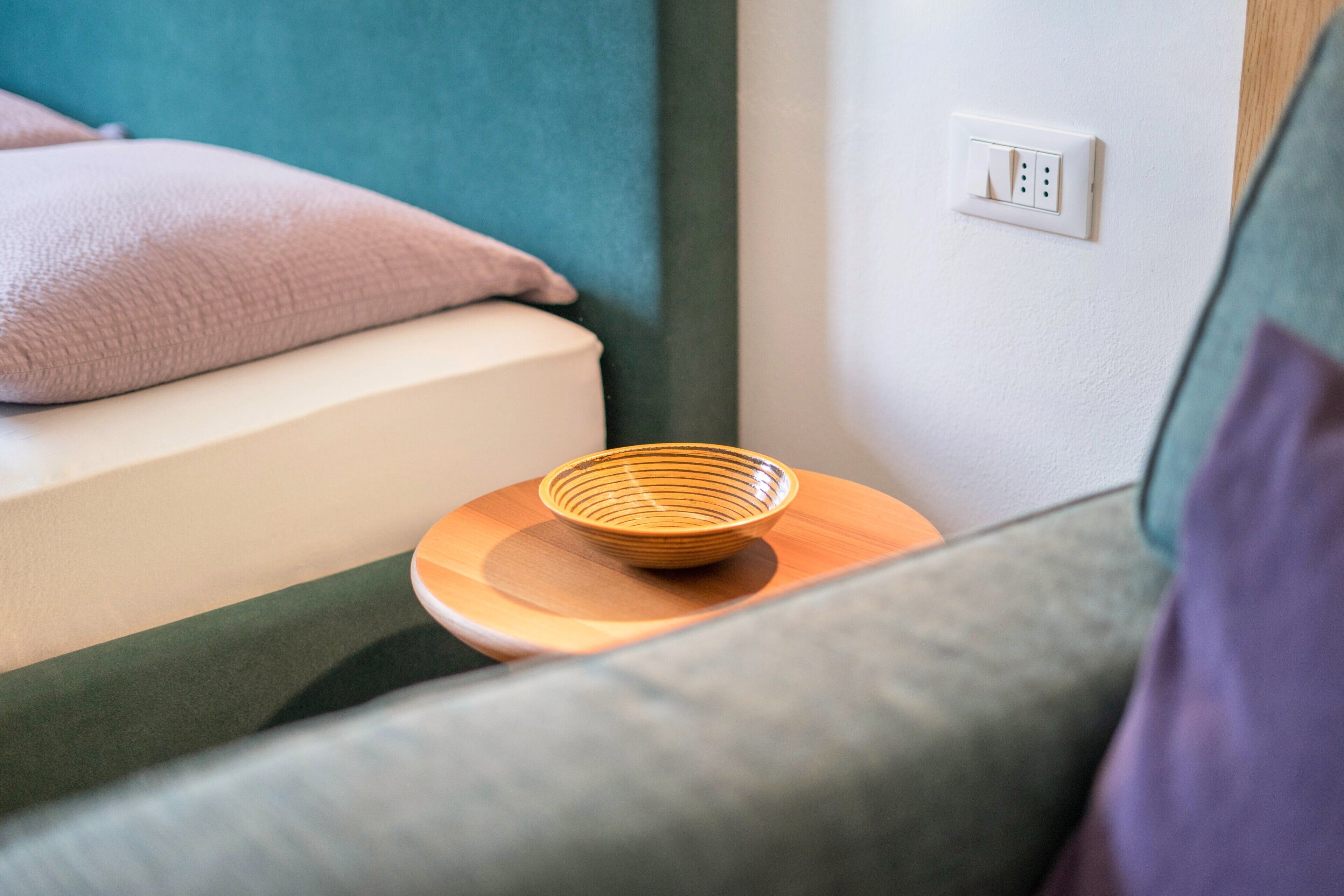 A room with a bed, a bedside table with a bowl, a wall socket, and a partial view of a couch.