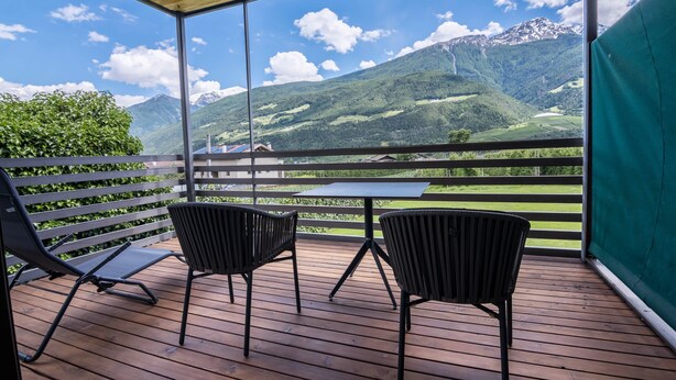 Uma varanda com piso de madeira, duas cadeiras pretas, uma mesa e vista para colinas e montanhas verdes.