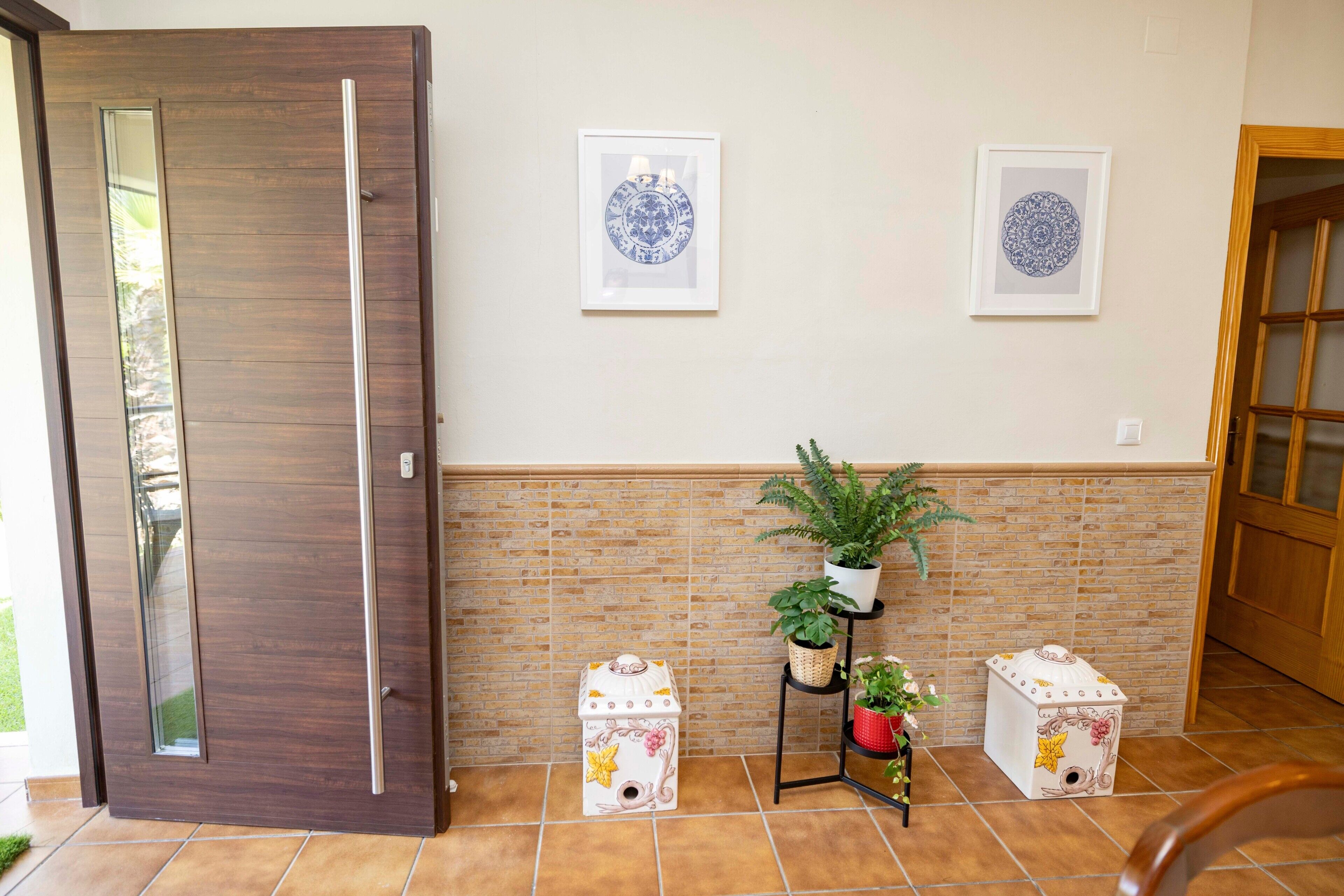 A room with a wooden door, tiled floor, two wall-mounted framed artworks, a potted plant on a shelf, and two decorative storage boxes.