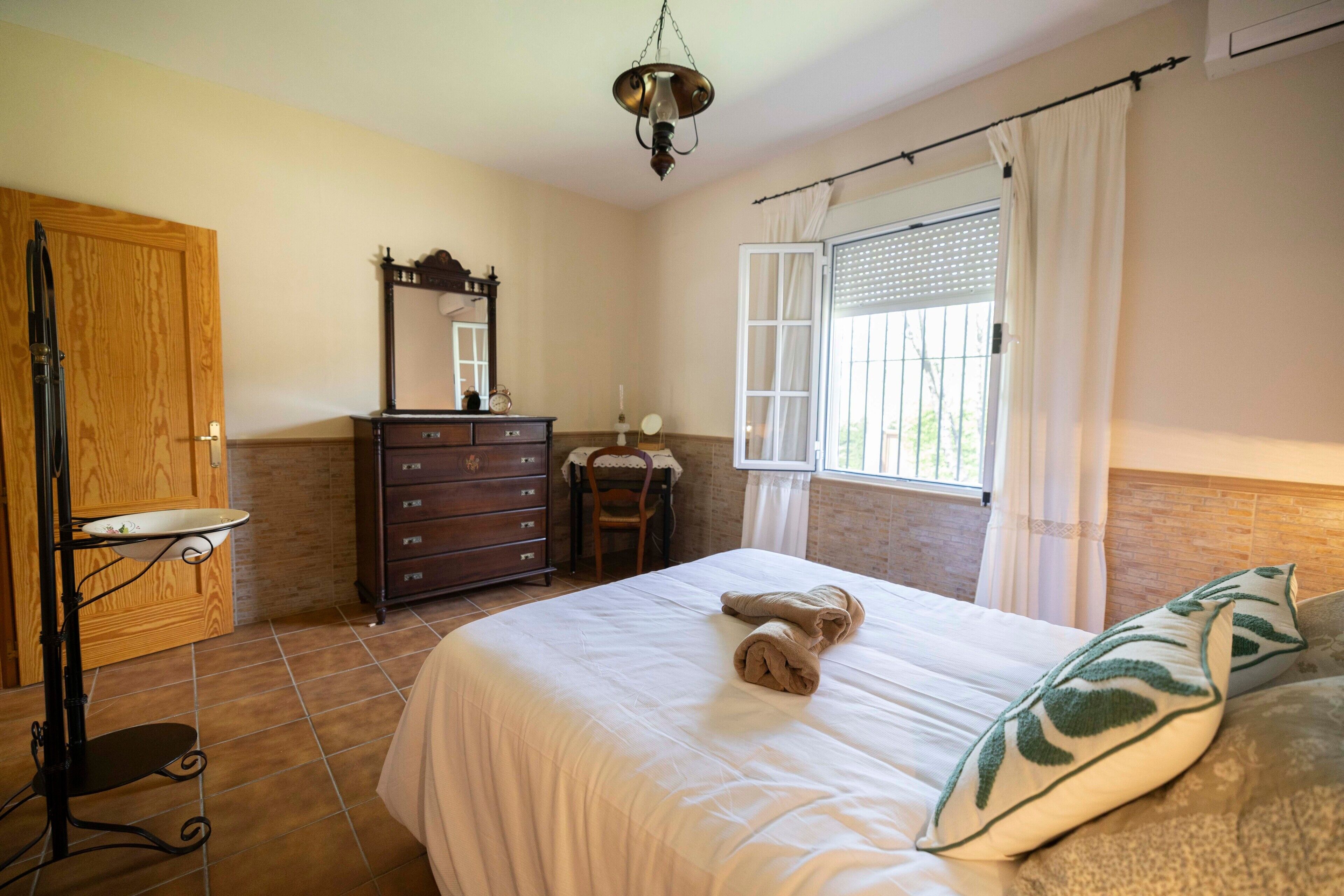 Une chambre à coucher comprenant un lit, une commode, un miroir, une fenêtre avec des rideaux et un luminaire suspendu.