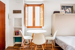 A room with a wooden door, a white round table with four chairs, a bed, a shelf with books, and a window with open shutters.