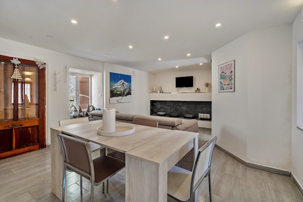 Un salon moderne avec une cheminée, un coin repas avec une table et des chaises en bois, et un téléviseur fixé au mur.