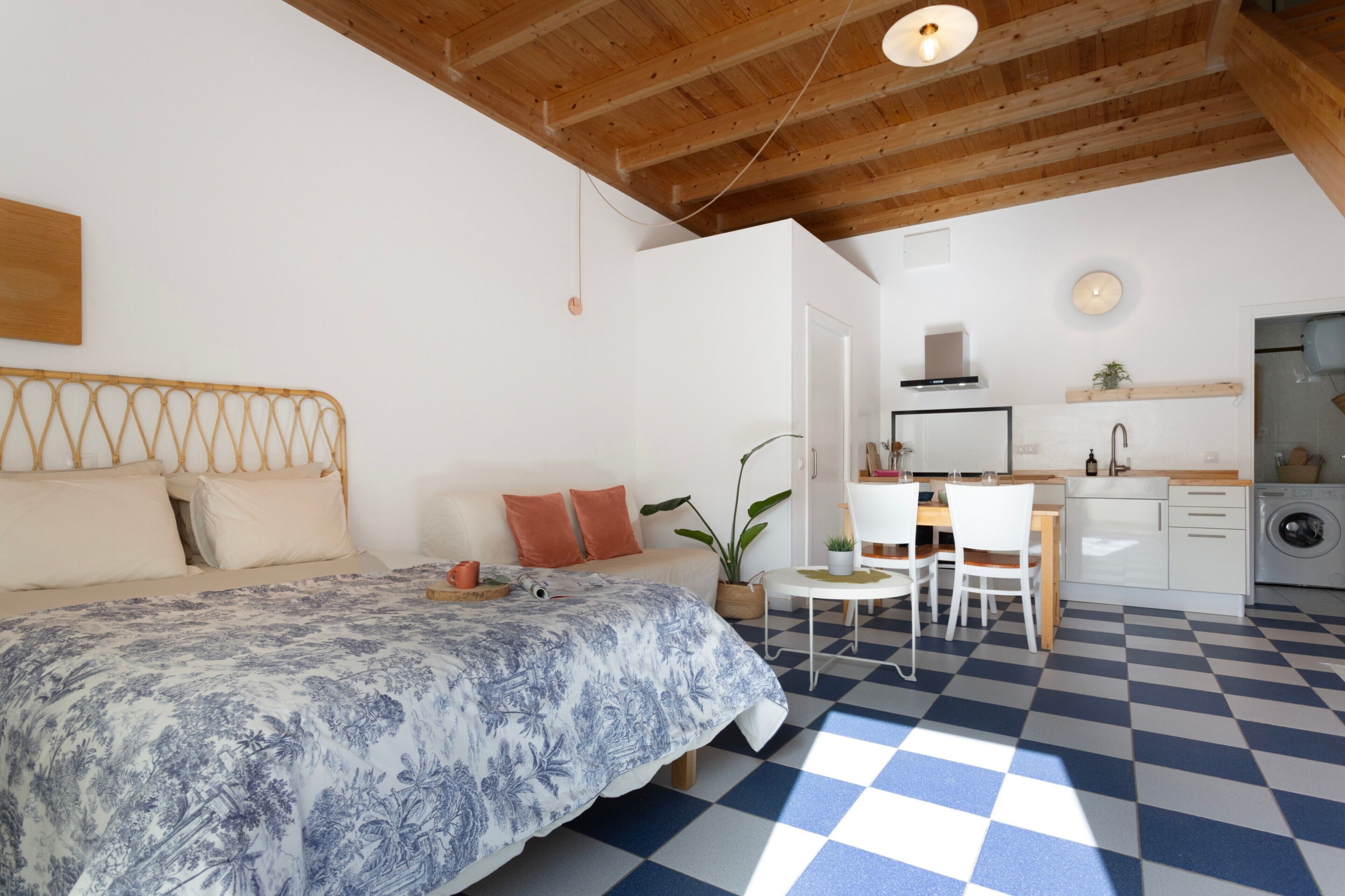 A bedroom with a checkered floor, a bed with a patterned bedspread, a wooden ceiling, and a small kitchen area with a sink and a washing machine.