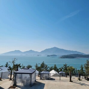 A coastal view with a clear sky, a body of water, and a distant mountain range. There are several tents and a fenced area with trees.
