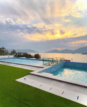 A swimming pool with a glass balustrade, surrounded by a well-maintained lawn and a scenic view of a lake and mountains during sunset.